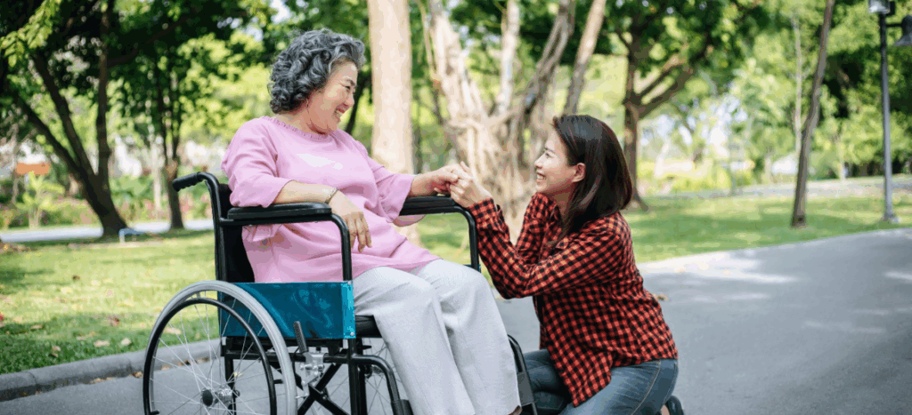 A young woman kneeling and holding hands with an elderly woman sitting in a wheelchair, sharing a warm moment together in a green park.
