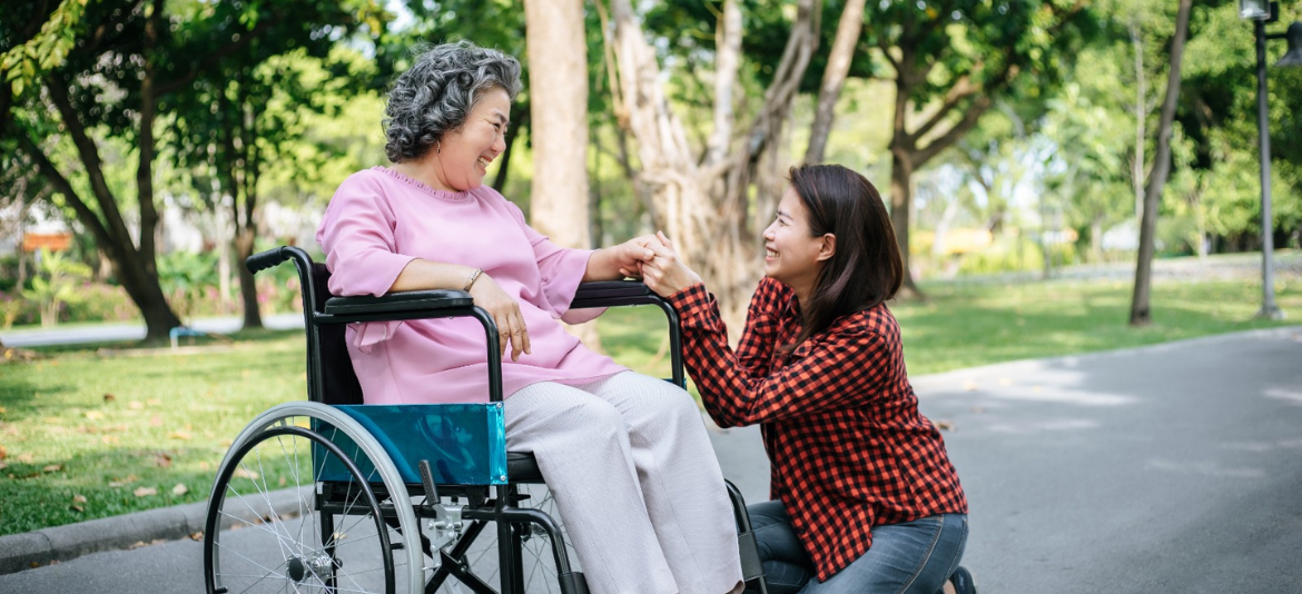 A young woman kneeling and holding hands with an elderly woman sitting in a wheelchair, sharing a warm moment together in a green park.