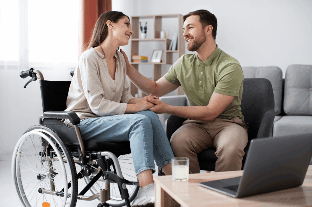 A man sits beside a woman using a wheelchair, holding her hands and smiling together in a comfortable living room.