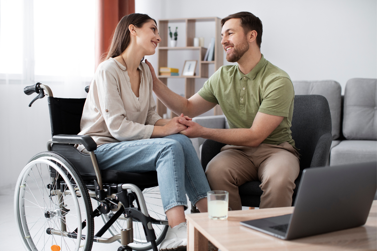 A man sits beside a woman using a wheelchair, holding her hands and smiling together in a comfortable living room.