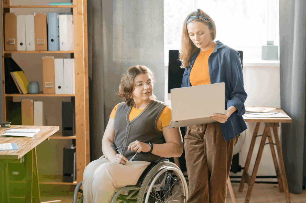 Two women working together in an office, one using a wheelchair while reviewing information on a laptop with a colleague