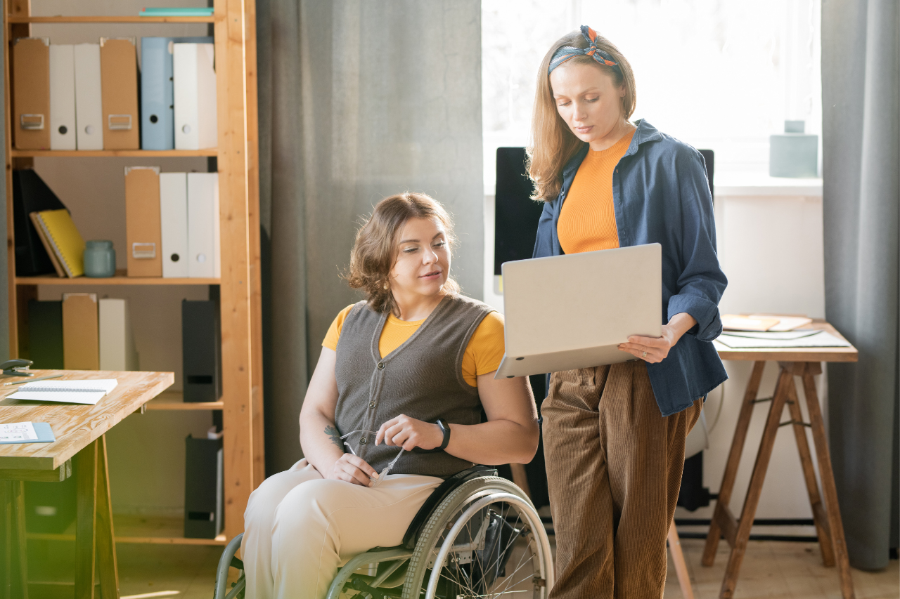 Two women working together in an office, one using a wheelchair while reviewing information on a laptop with a colleague